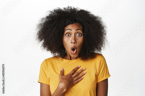 Enthusiastic Black woman looking surprised, staring impressed and fascinated at camera, standing in yellow t-shirt over white background