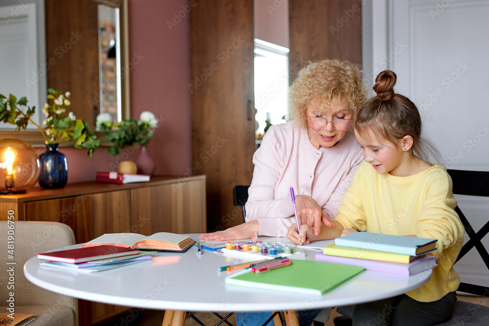 Caucasian aged woman spend time with daughter drawing and doing homework at home. Senior grandma happy relax with young girl sitting behind desk in living room, at daytime in cozy room Caucasian aged woman spend time with daughter drawing and doing homework at home. Senior grandma happy relax with young girl sitting behind desk in living room, at daytime in cozy room