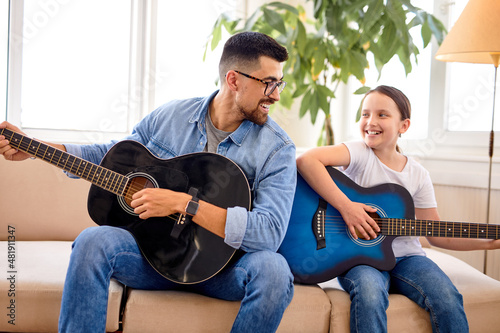 cute little girl and handsome father play guitar sitting on couch at home. father's day. care and education of children. side view on young caucasian family spending weekends at home, leisure time