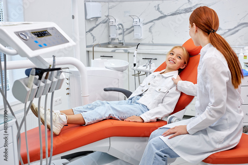 Happy little caucasian child girl in dental surgery waiting on examination couch, side view on confident female redhead dentist calm kid girl down, having nice talk. dental equipment in cabinet
