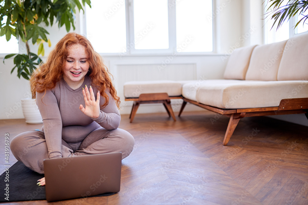 Happy chubby redhead woman preparing for an online fitness class, caucasian fat overweight female in sportstwear sit on mat using laptop, say hello. Distance training during the quarantine period Happy chubby redhead woman preparing for an online fitness class, caucasian fat overweight female in sportstwear sit on mat using laptop, say hello. Distance training during the quarantine period