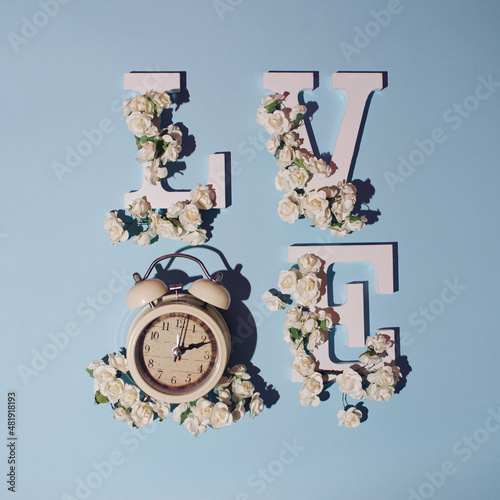 White letters LOVE on a pastel, soft blue background.  The letters are decorated with small white flowers and alarm clock. Message: Spring is coming,  nature is waking up, and it's time for tenderness