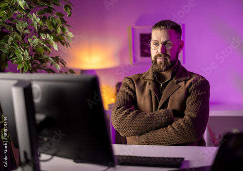 Caucasian bearded man sitting at desk with crossed arms and looking on pc monitor. Soft violet light at dark living room. Remote work and people concept.