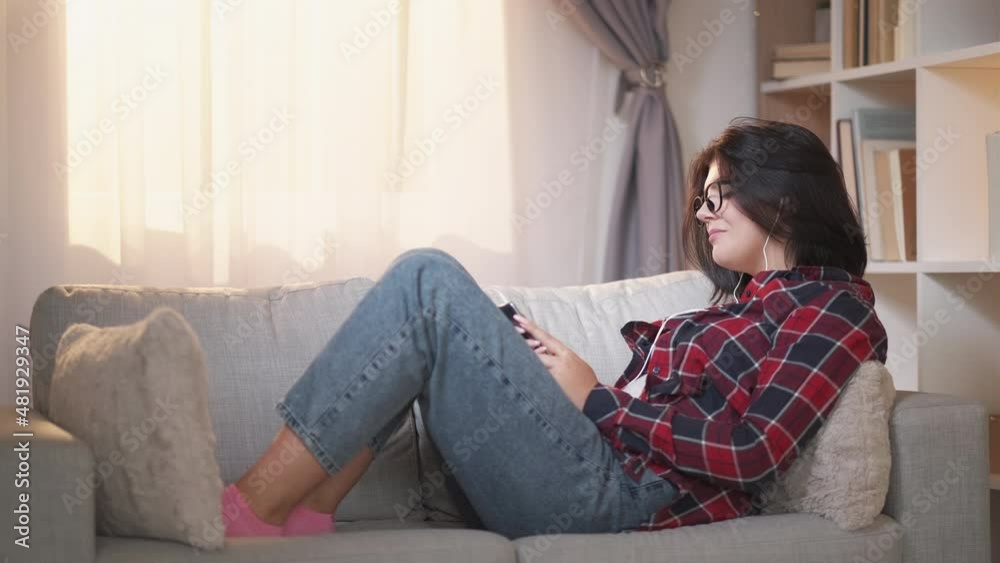 Mobile leisure. Gadget relaxing. Stay home. Cheerful millennial woman in earphones listening phone playlist music browsing internet on comfy couch at living room. Mobile leisure. Gadget relaxing. Stay home. Cheerful millennial woman in earphones listening phone playlist music browsing internet on comfy couch at living room.
