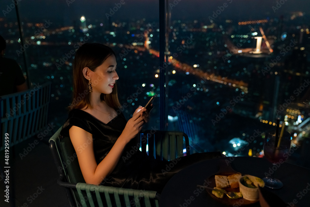 Beautiful Asian woman using mobile phone for social media or online shopping while waiting for meeting with her friends at skyscraper outdoor rooftop restaurant in the city at night.