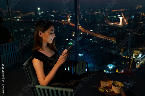 Beautiful Asian woman using mobile phone for social media or online shopping while waiting for meeting with her friends at skyscraper outdoor rooftop restaurant in the city at night.