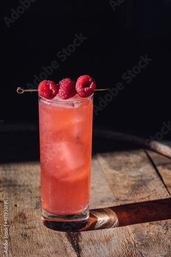 pink cocktail in tall Collins glass with ice and three raspberries on cocktail pick photo