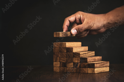 Close-up hand is placing wood block tower stacked in pyramid stair step with caution to prevent collapse or crash concepts of financial risk management and strategic planning.