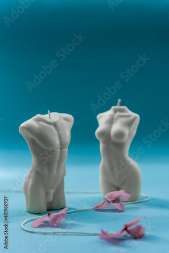 Man and woman bodies made of a wax. Romantic shapes for candles. Close up on two aroma candles. Flowers and plants around the candles. Handmade soy wax candles on the green background.