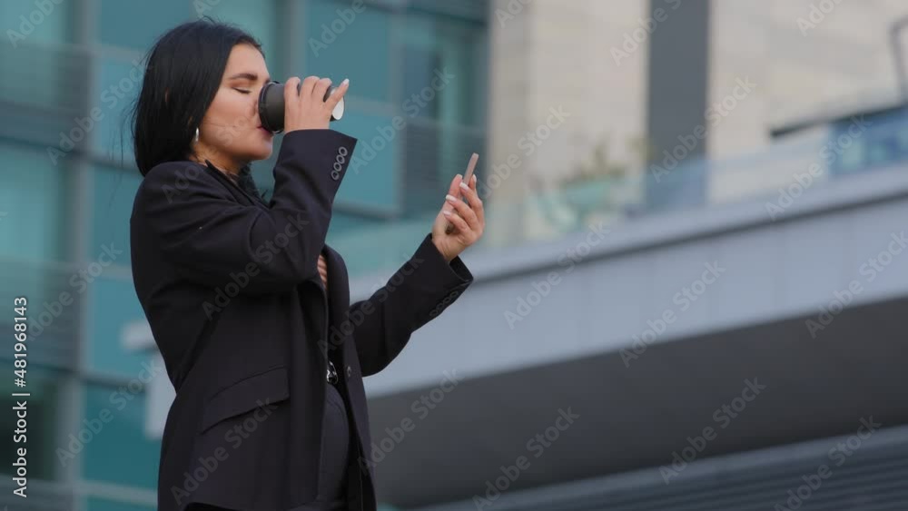 Young hispanic businesswoman walking street background of office building checks email on phone uses mobile app makes purchase online sends text message on smartphone drinking coffee from paper cup Young hispanic businesswoman walking street background of office building checks email on phone uses mobile app makes purchase online sends text message on smartphone drinking coffee from paper cup