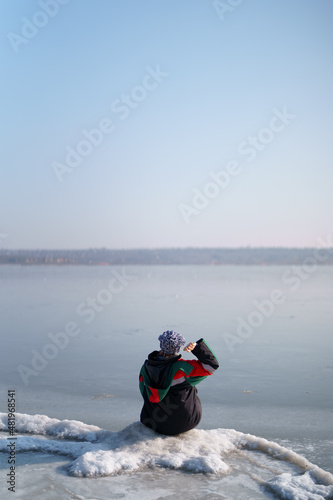 A woman in a colorful thermal costume enjoying winter while sitting and watching a frozen lake with blue sky in the background. Freedom