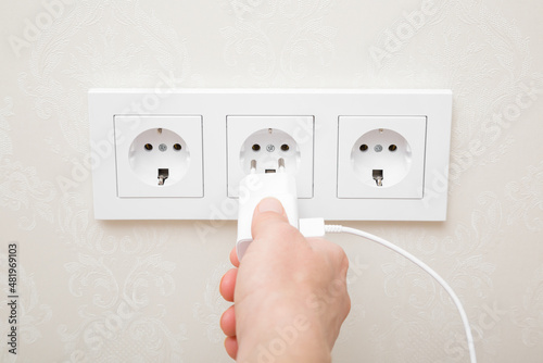 Young adult woman hand holding and plugging white electrical plug for smartphone charging in wall outlet socket at home. Closeup. Point of view shot.