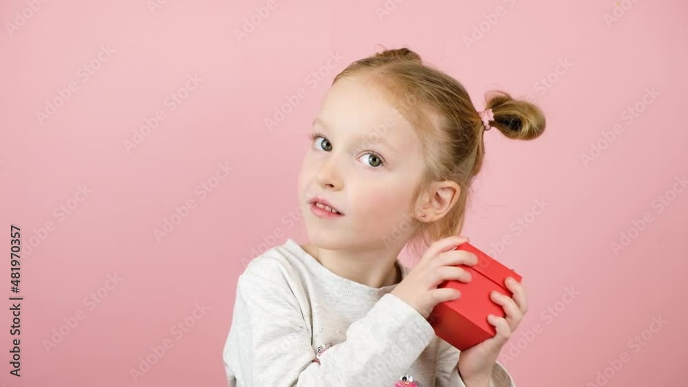 Funny curious little blonde girl smiling and shaking the red gift box on pink background. Valentines Day or Mothers Day concept Funny curious little blonde girl smiling and shaking the red gift box on pink background. Valentines Day or Mothers Day concept