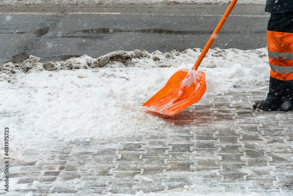 Removing snow from the sidewalk after snowstorm. A road worker with a shovel in his hands and in special clothes cleans the sidewalk and the road from snow. Snowstorm and hurricane in the city. Removing snow from the sidewalk after snowstorm. A road worker with a shovel in his hands and in special clothes cleans the sidewalk and the road from snow. Snowstorm and hurricane in the city.