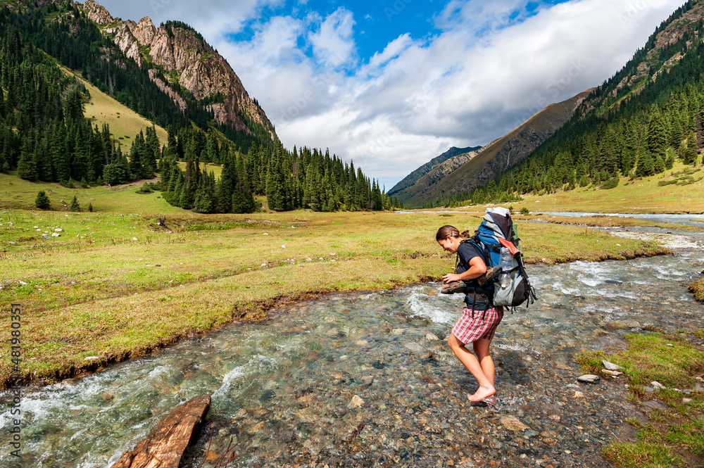 Fototapeta premium Young woman with big backpack barefoot crossing river. Shoeless female backapcker in river. Karakol valley, Issyk-kul region, Ala-kul lake Terskey Alatau mountain range, Kyrgyzstan, Central Asia.