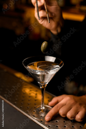 focus on green berry olive over transparent martini glass with cocktail on bar counter