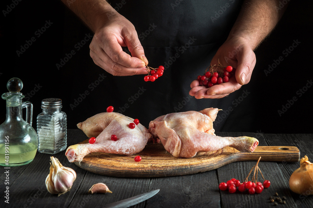 Professional chef prepares raw chicken legs in the restaurant kitchen. The cook puts the red viburnum on chicken leg before baking. National dish