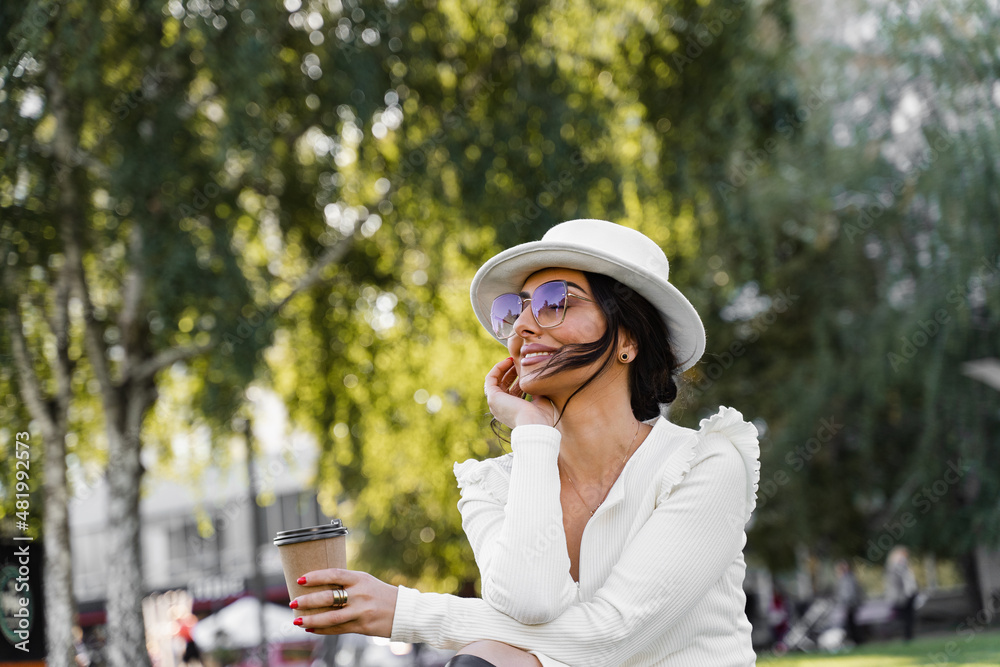 Fashion woman enjoy coffee. Attractive model weared white dress, hat and sunglasses posing on the street.