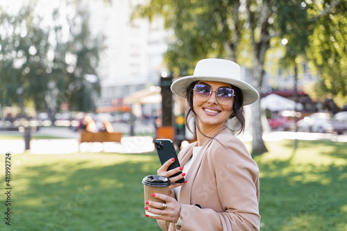 Business woman with mobile phone and cup of coffee on the street. Attractive girl weared official white dress, jacket and hat.