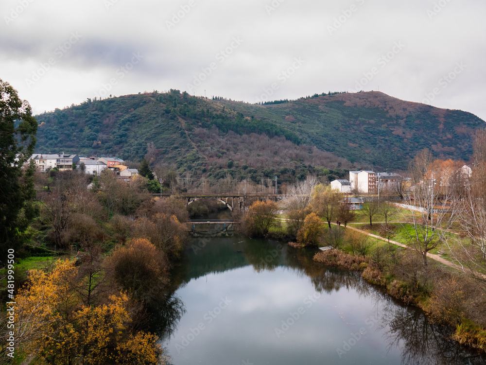 View of the Sil river as it passes through the city of Ponferrada with vegetation around it and the train tracks on a bridge and the Pajariel in the background on a cloudy day