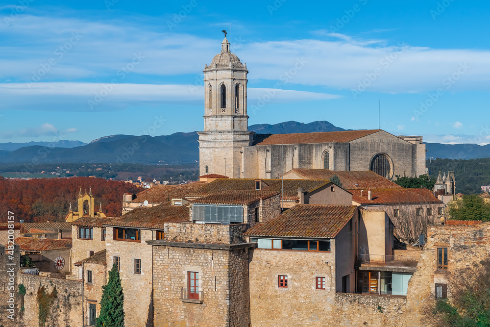 Girona Cathedral rises above the tiled roofs of the Gothic Quarter in the Old Town. Medieval cityscape with ancient buildings against the backdrop of mountains and blue sky on a sunny autumn day