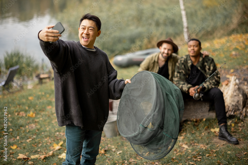 Asian man taking selfie on smartphone with multiracial male friends. Men resting and spending time together on river in nature at autumn. Concept of leisure, weekend and vacation. Idea of friendship Asian man taking selfie on smartphone with multiracial male friends. Men resting and spending time together on river in nature at autumn. Concept of leisure, weekend and vacation. Idea of friendship