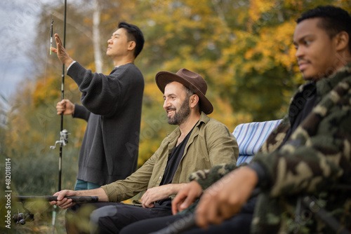 Multiracial male friends resting, talking and fishing on river or lake coast. Concept of leisure, hobby and weekend in nature. Idea of friendship and spending time together. Autumn day