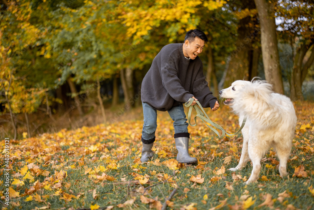 Smiling asian man playing with Maremmano-Abruzzese Sheepdog on river or lake coast at autumn morning. Concept of rest and weekend in nature. Adult male wearing boots and warm clothes