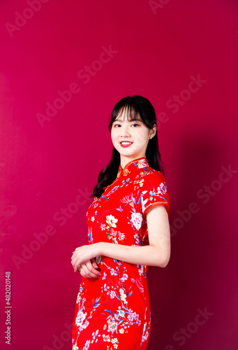 Smiling asian young woman in traditinal red cheongsam dress pointing hand to empty space on red background.