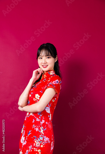 Smiling asian young woman in traditinal red cheongsam dress pointing hand to empty space on red background.