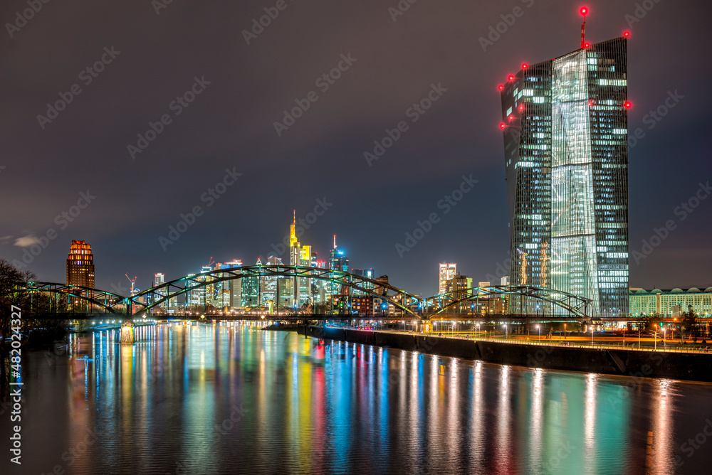 Die EZB (Europäische Zentralbank) in Frankfurt am Main vor dem Nachthimmel und der beleuchteten Skyline mit dem Fluß Main mit Lichtreflexen im Vordergrund