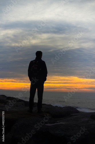 Beautiful sunset on the seashore. A man standing with a backpack on a rock by the sea against a colorful sunset sky.