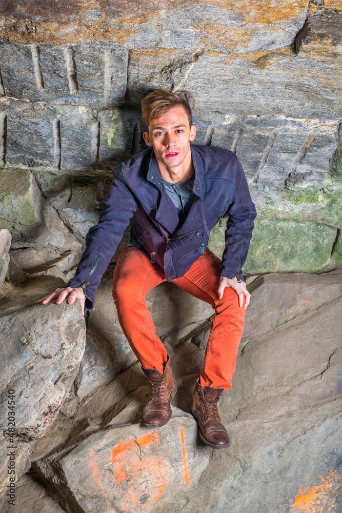 Bending over his body, frowned, a young guy is standing in a cave of rocks, looking up, hair messed, distressful. .