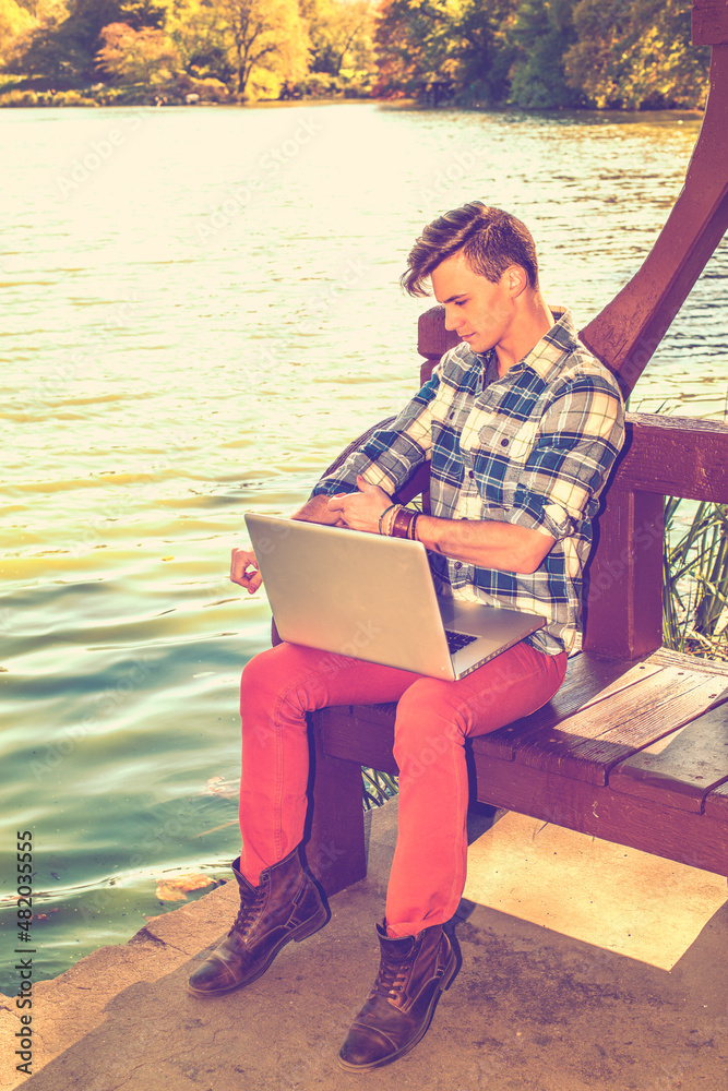 Dressing in a long sleeve, patterned shirt, red pants, brown boots, rolling over sleeves, one young guy is working on a laptop computer by a lake in afternoon. Retro filtered effect..