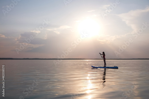 Silhouette of a beautiful woman swimming on big lake staying Paddle SUP Board at sunset background. © gleaminvisible