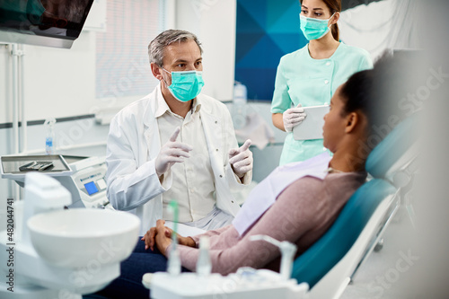 Mature dentist with face mask talks to black female patient at dentist's office.