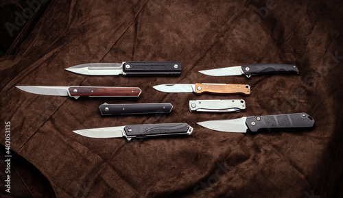 A variety of folding and pocket knives lie on brown fabric. A versatile pocket tool and self-defense tool.