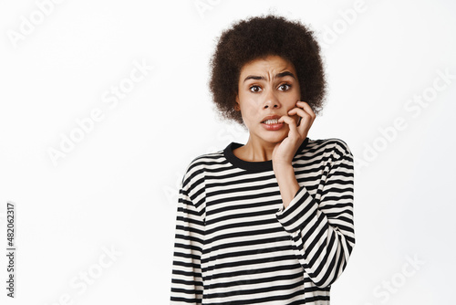 Portrait of scared, anxious black girl biting fingers and looking worried at camera, frightened of something, standing over white background