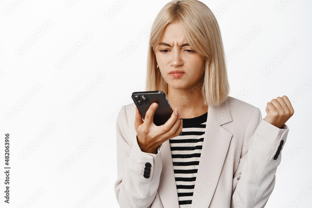 Angry corporate woman, business girl looking at mobile phone with furious face and frowning grimace, standing over white background Angry corporate woman, business girl looking at mobile phone with furious face and frowning grimace, standing over white background