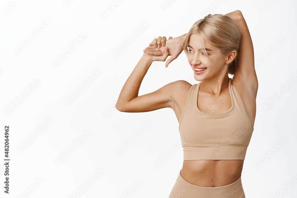 Young attractive fitness girl, female athlete stretching arms and warm up for jogging, runner doing exercises, workout in gym, showing fit strong body and biceps, white background Young attractive fitness girl, female athlete stretching arms and warm up for jogging, runner doing exercises, workout in gym, showing fit strong body and biceps, white background