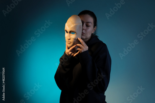 Hiding behind a mask, a young woman in a dark hoodie hides her face with a mask, self-identification problems and impostor syndrome. Portrait in the studio on a dark gray background.