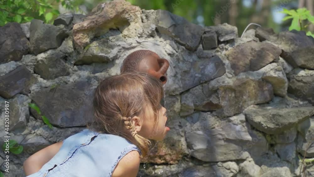 Little girl drinks water from a pure spring in the forest in the mountains. Kid drinking fresh and cool spring flows from the ground. Cool and delicious spring water. Natural underground water source Little girl drinks water from a pure spring in the forest in the mountains. Kid drinking fresh and cool spring flows from the ground. Cool and delicious spring water. Natural underground water source