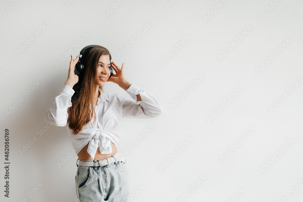 Cheerful smiling young brunette in white shirt and jeans, listening to music in blalck headphones, over white studio. Girl listening to music in headphones, dancing Cheerful smiling young brunette in white shirt and jeans, listening to music in blalck headphones, over white studio. Girl listening to music in headphones, dancing