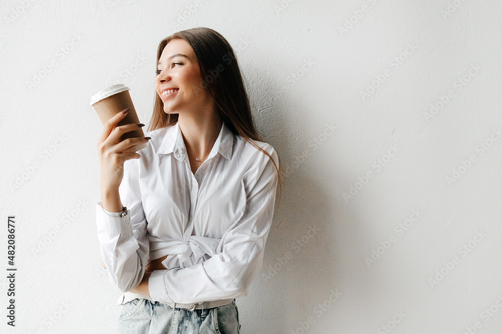 Young beautiful brunette woman in white t-shirt and jeans, holding takeaway coffee cup, smiling and looking to the side over white background Young beautiful brunette woman in white t-shirt and jeans, holding takeaway coffee cup, smiling and looking to the side over white background