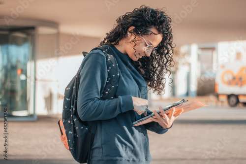 Muchacha Estudiante de cabello rizado oscuro estudiando y revisando el su teléfono móvil los apuntes del instituto en la entrada de su centro escolar antes de asistir al aula de estudios