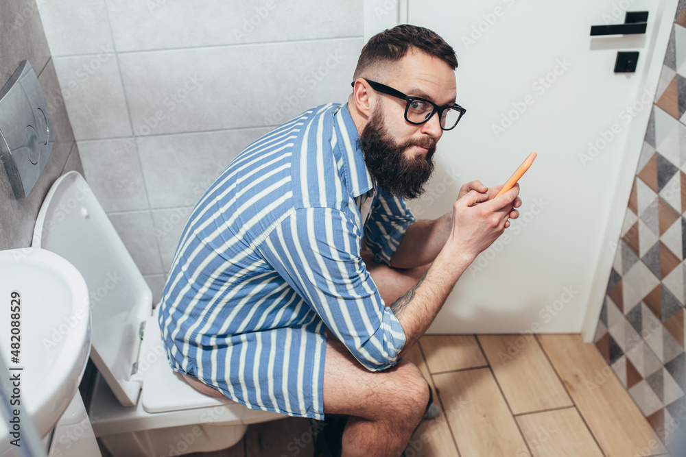 Handsome hipster guy with smartphone sitting on the toilet in the bathroom