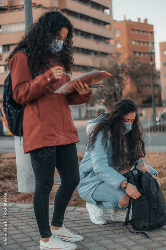 Jovenes niñas alumnas con mascarilla recogiendo sus apuntes con la mochila de la universidad mientras su compañera estudia los apuntes 