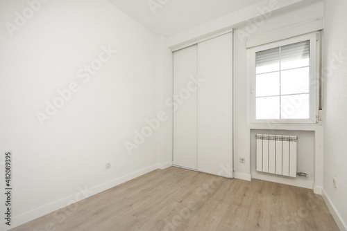 Empty room with light wooden floor, white wooden sliding wardrobe doors and aluminum window above aluminum radiator
