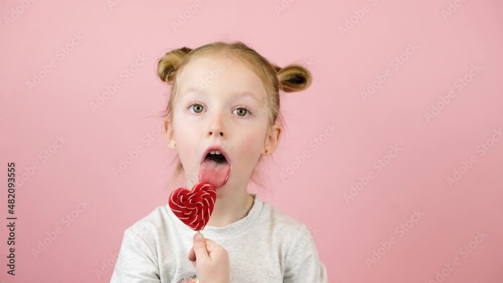 Funny little blonde girl smiling and licking red heart shape lollipop caramel on pink background. Valentines Day or Mothers Day concept Funny little blonde girl smiling and licking red heart shape lollipop caramel on pink background. Valentines Day or Mothers Day concept