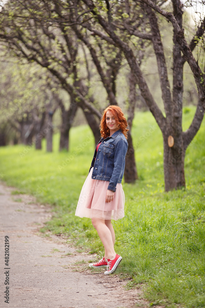 Active and happy woman in spring park. Young redhaired girl on background of green trees lane. Redhaired girl in peach skirt and jeans jacket and sneakers walks in nature. Young early spring